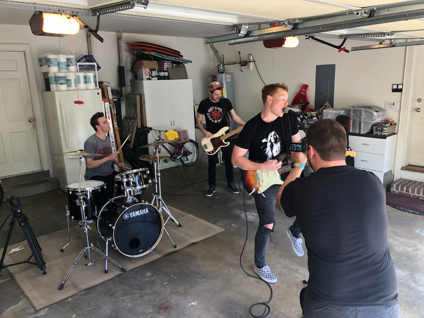 Behind-the-scenes of a band content shoot — drummer, bassist, and vocalist performing in a garage while a videographer films on a smartphone